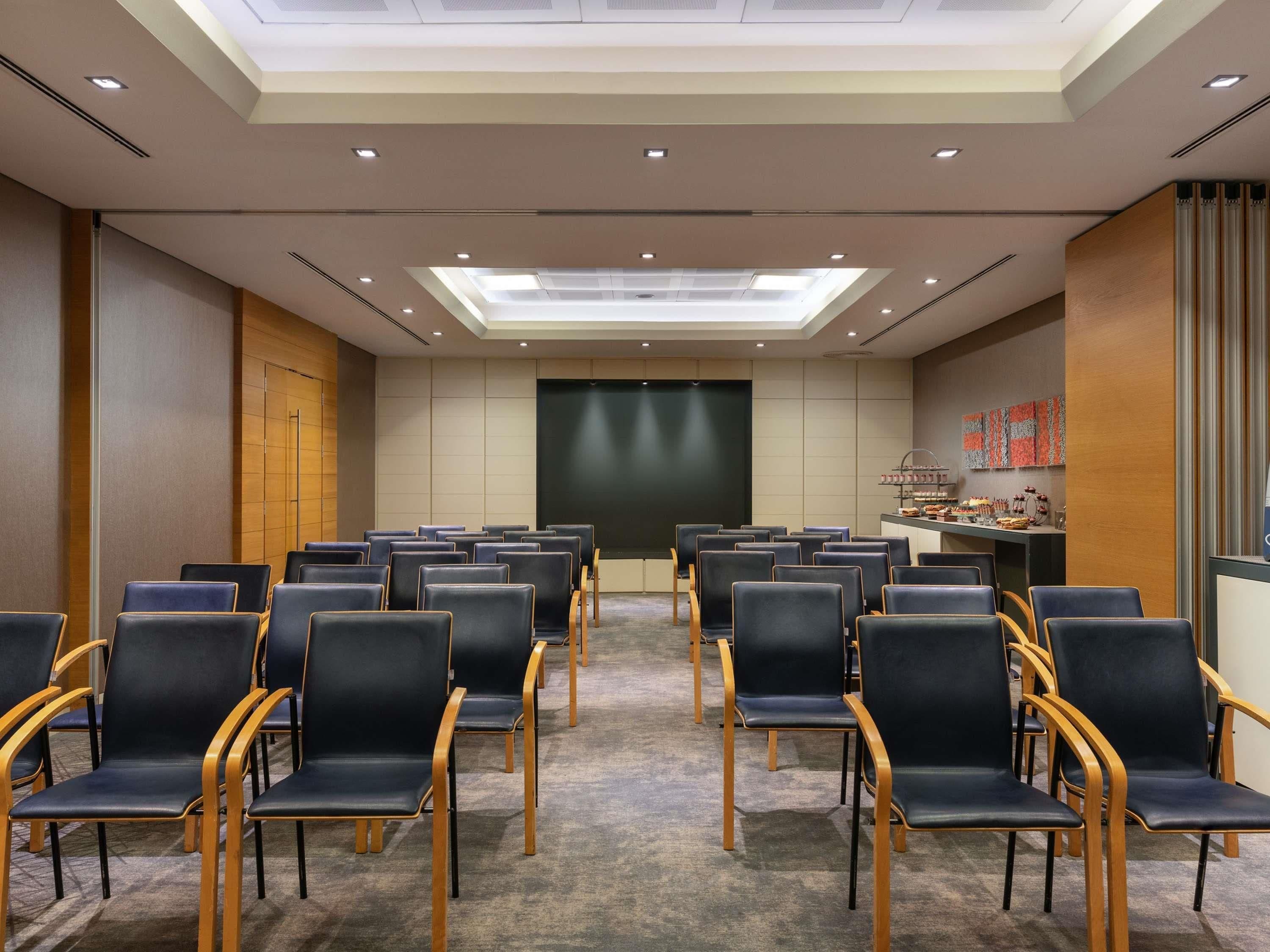 Un salón de conferencias con filas de sillas, una pantalla de proyector y una zona de bufé.