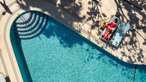 Una grande piscina azzurra e trasparente con bordo curvo, circondata da una terrazza piastrellata e lettini da relax.
