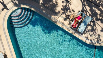 A large, clear blue swimming pool with a curved edge, surrounded by a tiled deck and lounge chairs.
