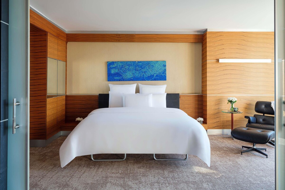 Ein modernes Hotelzimmer mit einem großen Bett, einer Wand im Holzdesign, einem blauen Kunstwerk, einem kleinen Tisch mit einer Vase und einem Ledersessel.