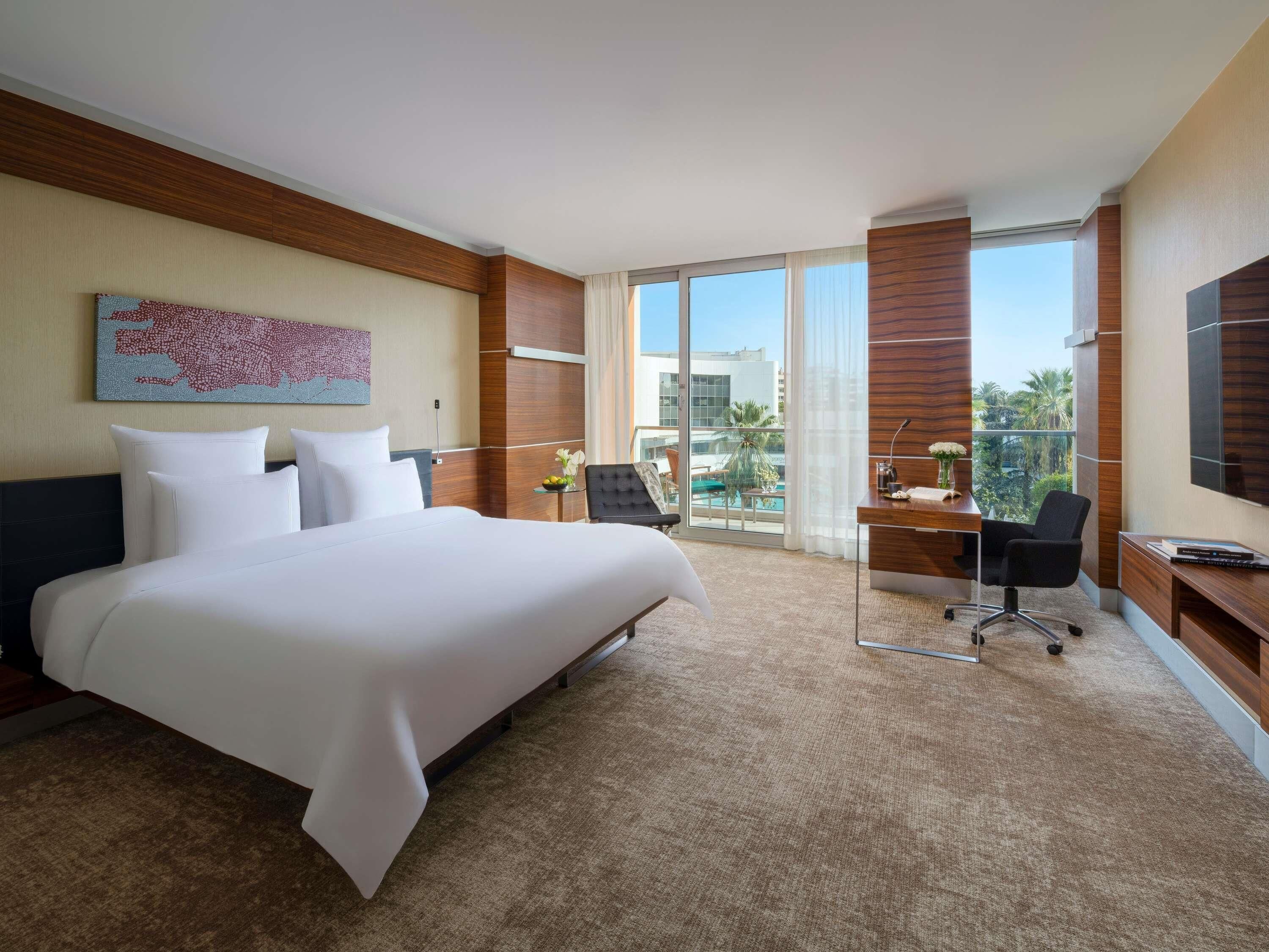 Habitación de hotel con una cama grande, un escritorio, una silla, un televisor y vistas a una piscina y edificios.
