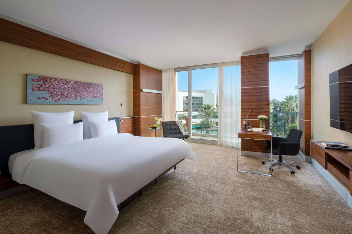 Une chambre d’hôtel avec un grand lit, un bureau, une chaise, une télévision et une vue sur une piscine et des immeubles.