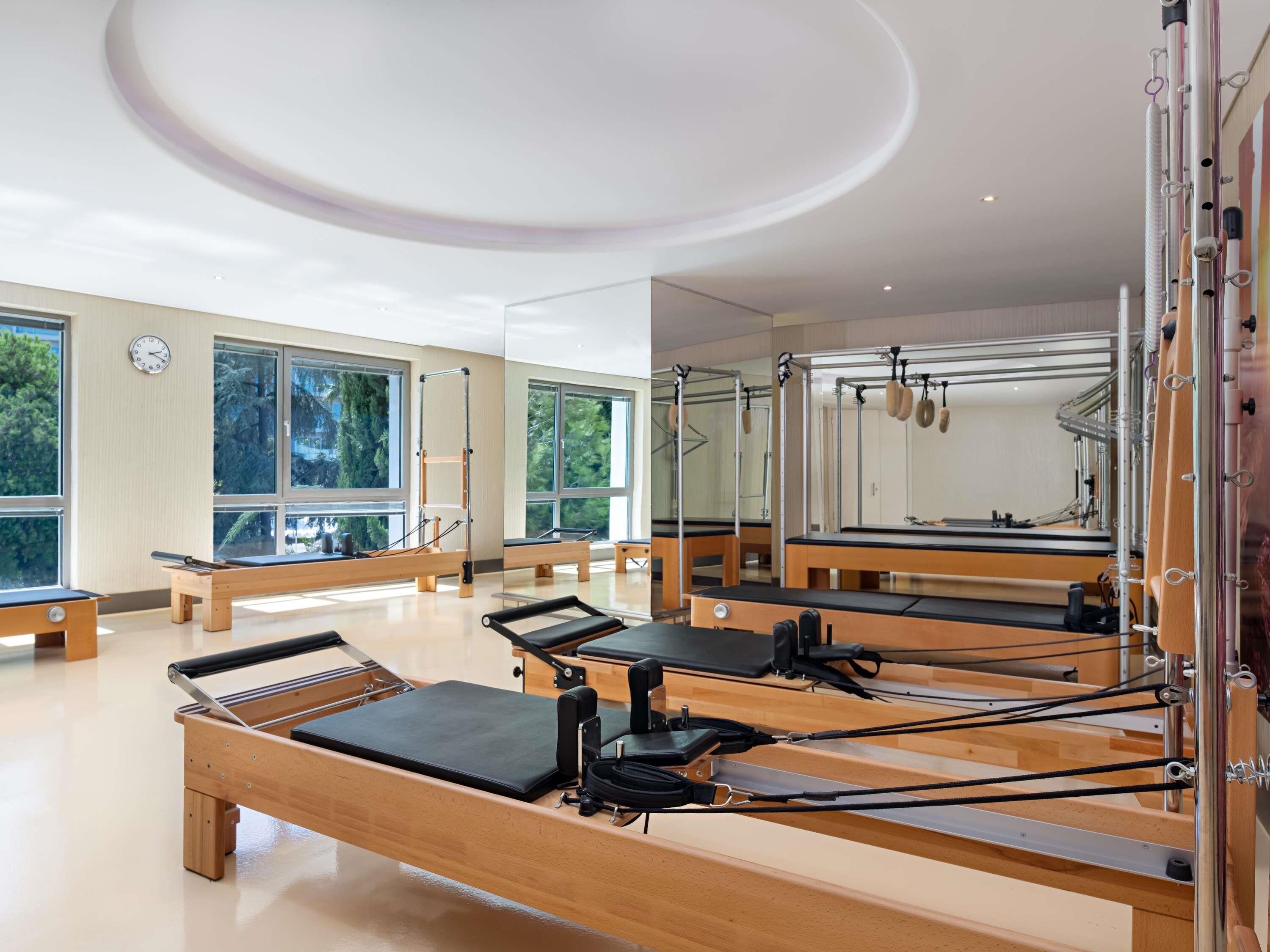 Un estudio de Pilates con máquinas reformadoras de madera, ventanas amplias y un reloj en la pared.