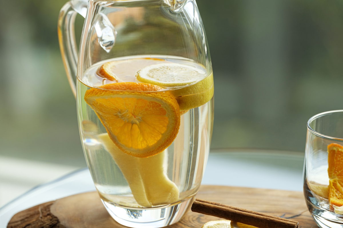 A clear glass pitcher with slices of lemon and orange, placed on a wooden tray.
