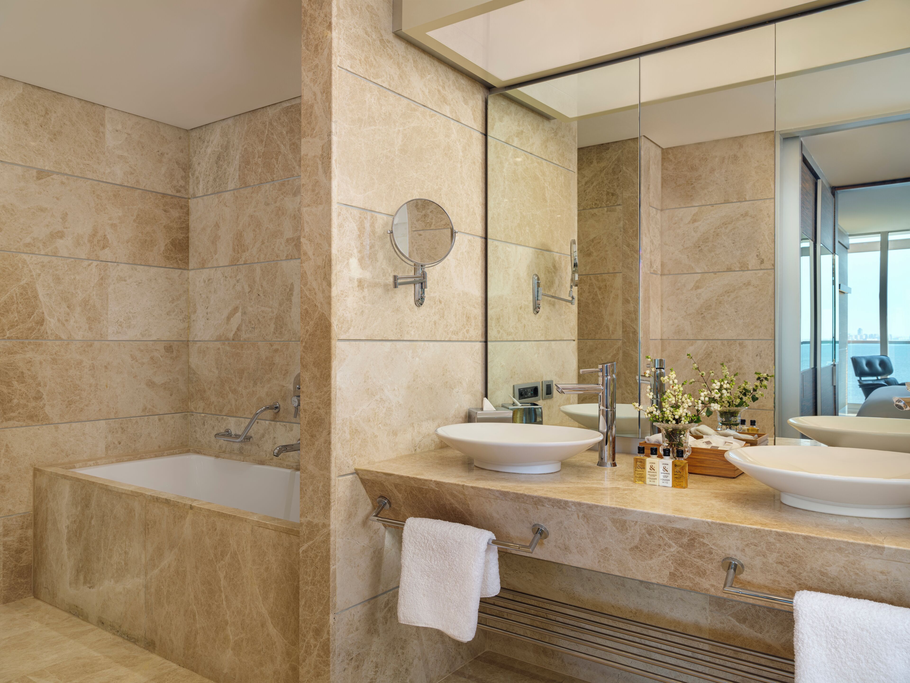 Une salle de bain moderne avec une baignoire, deux lavabos, un grand miroir et une vue sur la ville par la fenêtre.