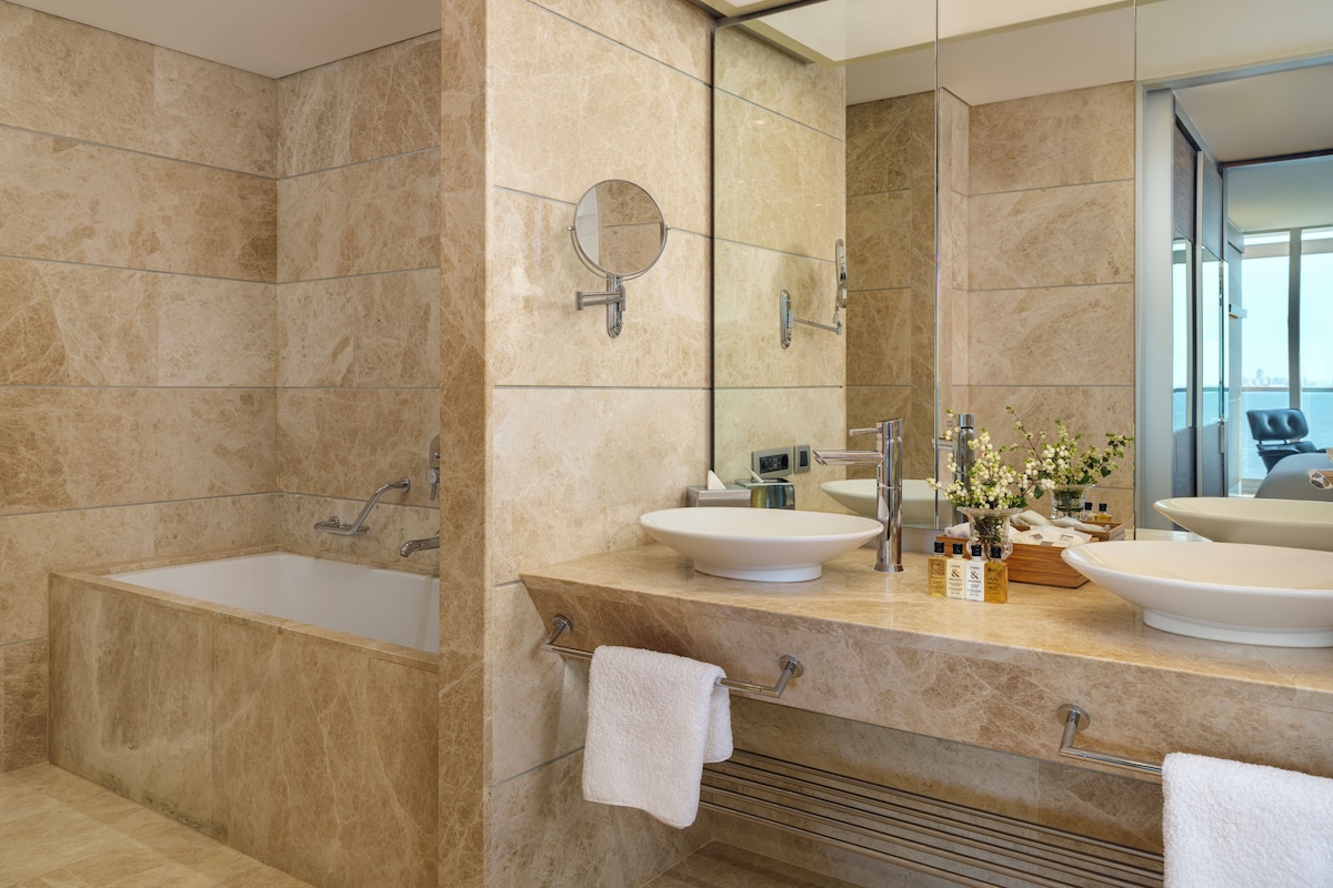 A modern bathroom with a bathtub, two sinks, a large mirror, and a view of the city through the window.