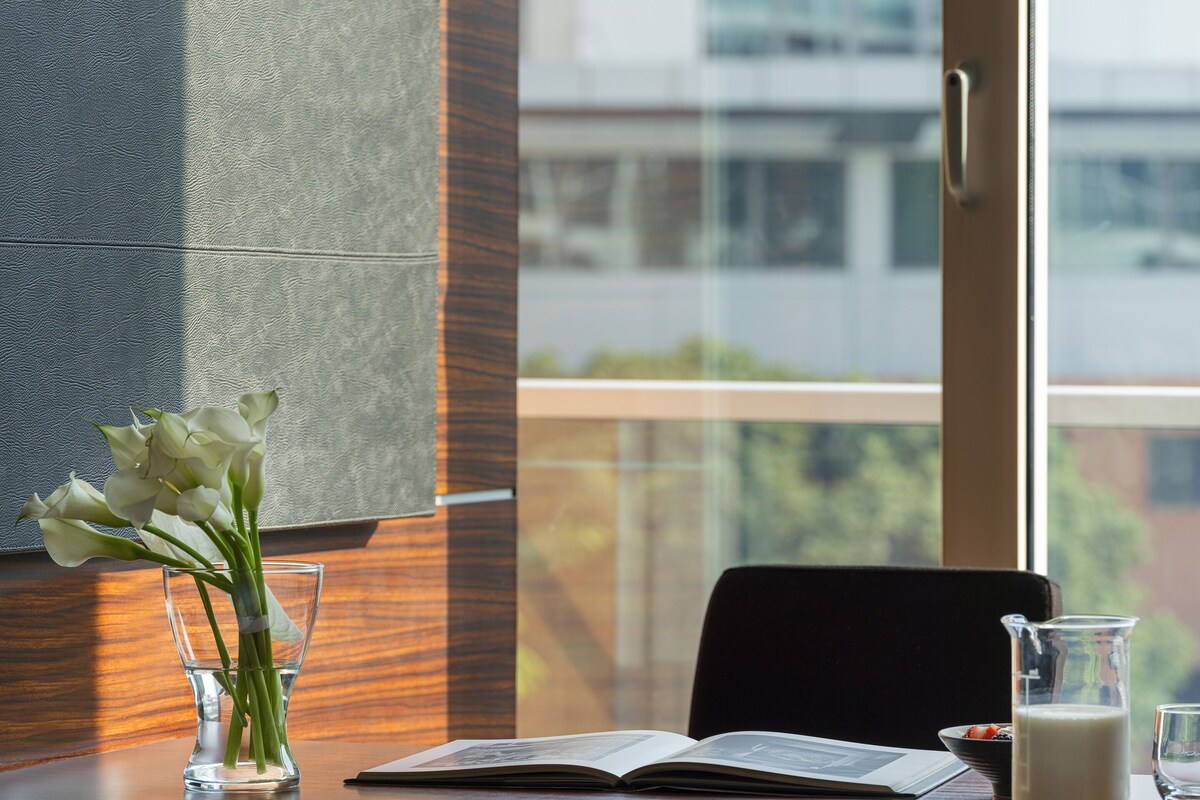 A modern office space with a wooden desk, a clear glass vase with white flowers, an open book, a glass pitcher of milk, and a black chair.
