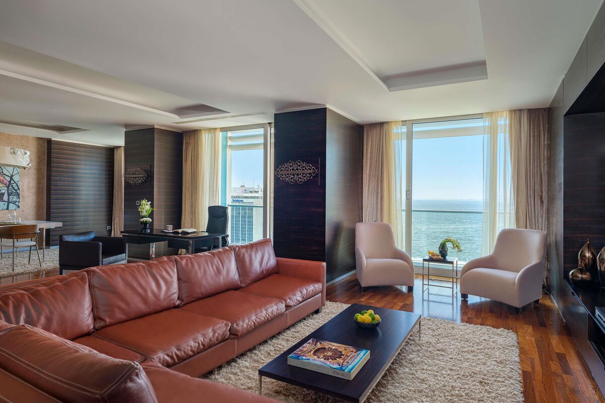 Ein modernes Wohnzimmer mit Ledersofa, Couchtisch und zwei Sessel. Ein Essbereich mit Tisch und Stühlen ist ebenfalls zu sehen. Große Fenster bieten Blick auf das Meer.