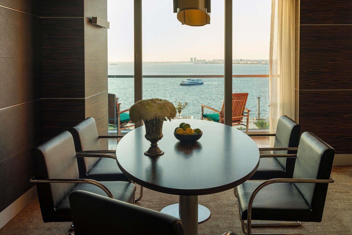 Une pièce moderne avec une table ronde, deux chaises, un vase de fleurs et un saladier rempli de fruits. Une grande fenêtre offre une vue sur la mer et sur le paysage urbain au loin.