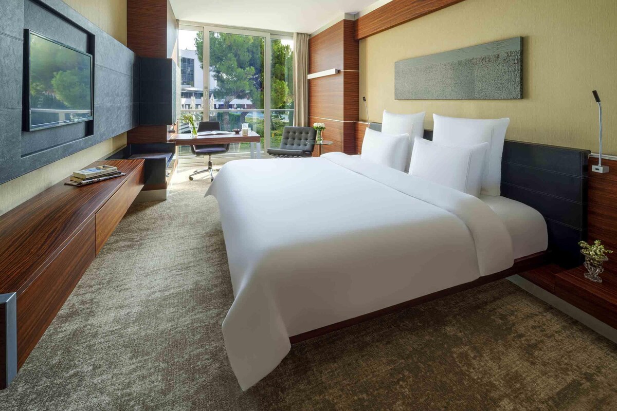Une chambre d’hôtel avec un grand lit, un bureau, une chaise, une télévision et une fenêtre donnant sur un paysage verdoyant.