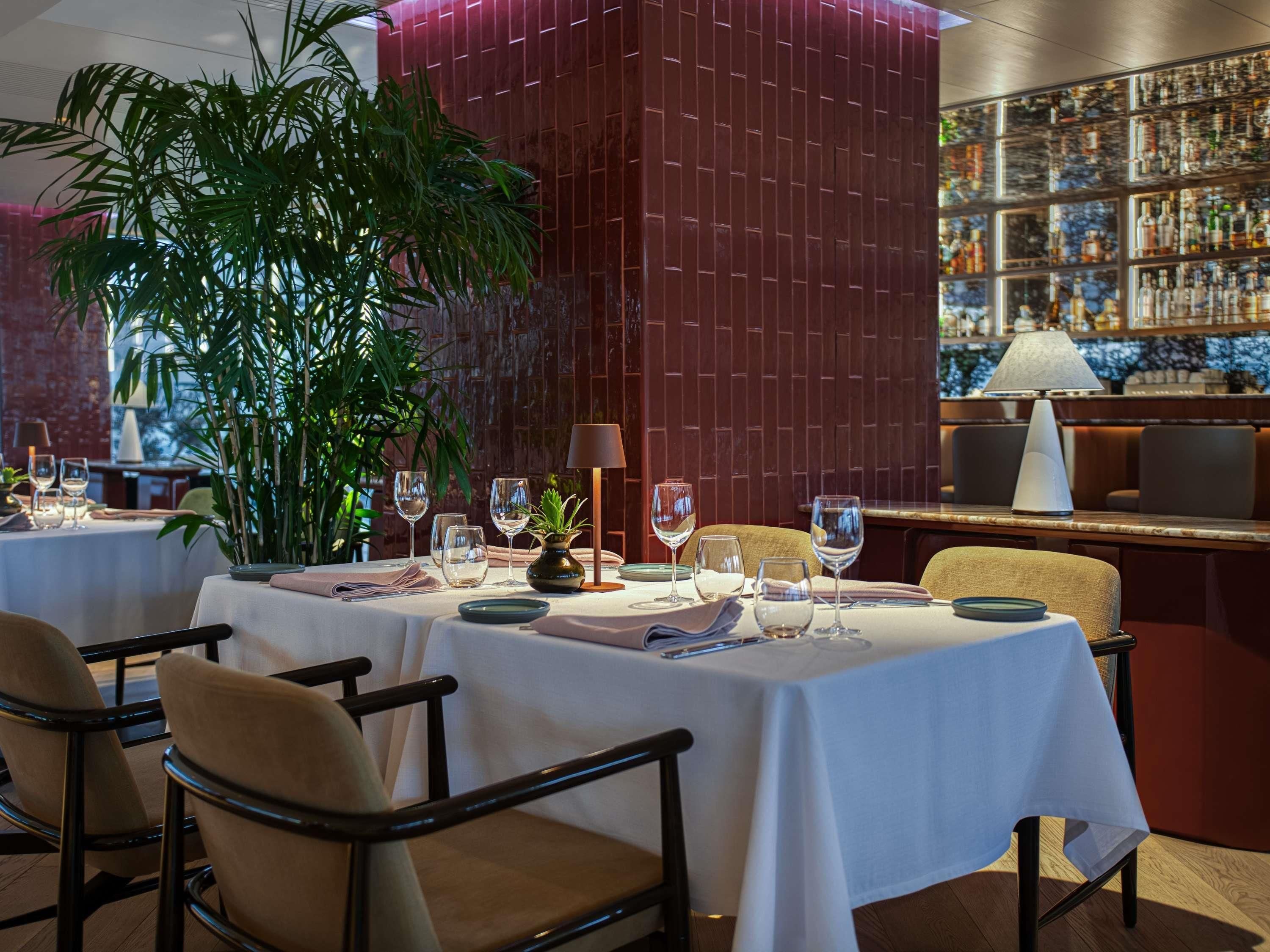 Ein Restaurantinneres mit gedeckten Tischen, einem großen Topfpflanzen und einer Wand mit Spirituosenflaschen.