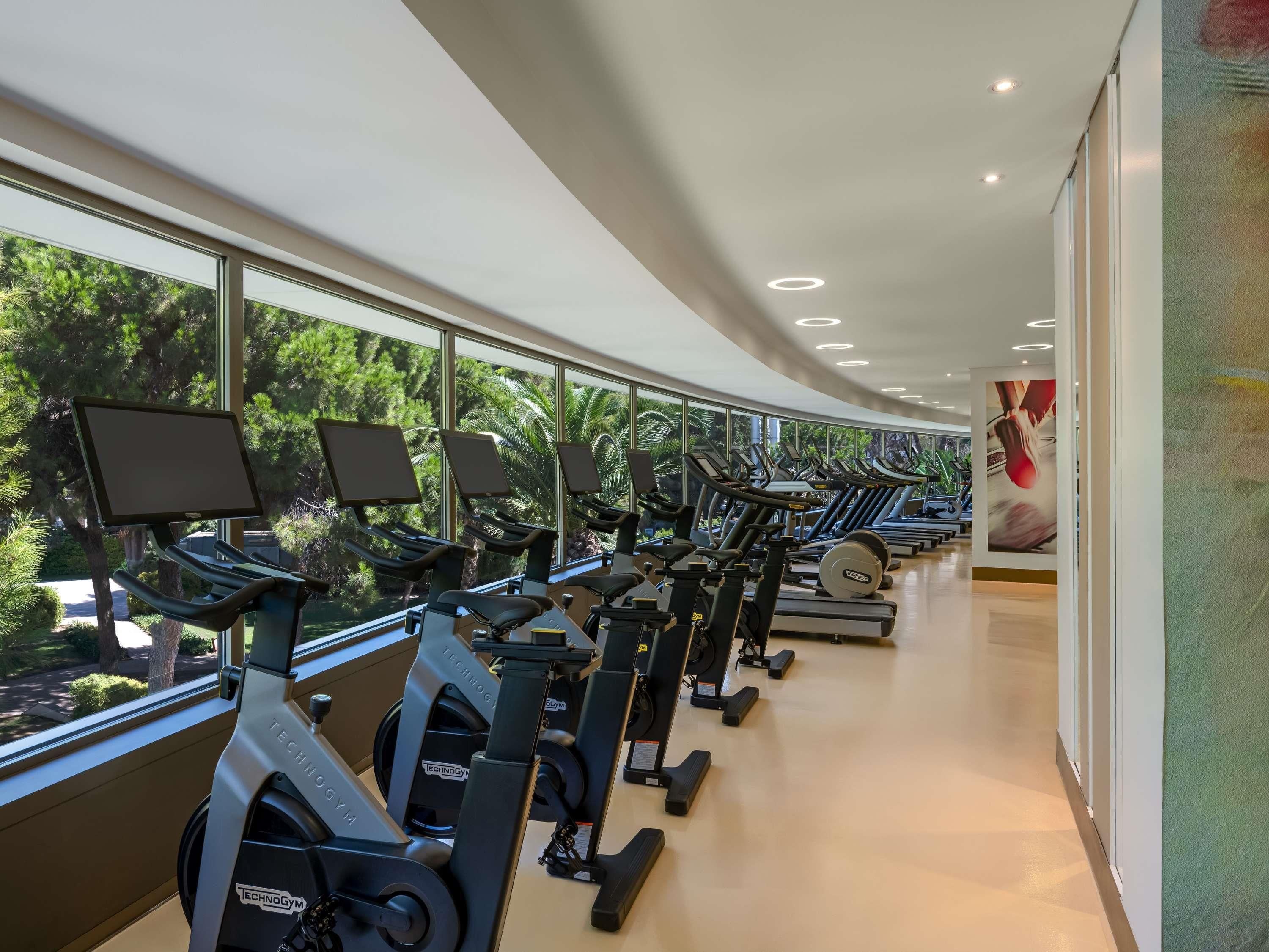 Une salle de sport équipée de vélos d’appartement et de tapis de course, de grandes fenêtres offrant une vue sur les arbres, et un aménagement propre et moderne.
