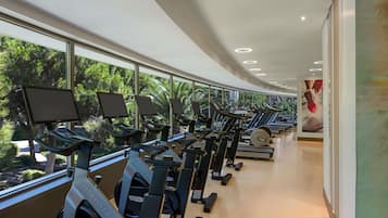 Un gimnasio con bicicletas estáticas y cintas de correr, amplias ventanas con vista a árboles, y un diseño moderno y cuidado.