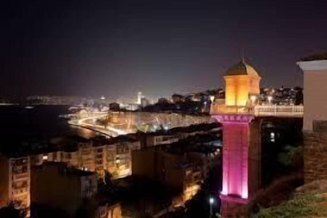 Vista nocturna de una ciudad costera con una torre destacada y un puente.