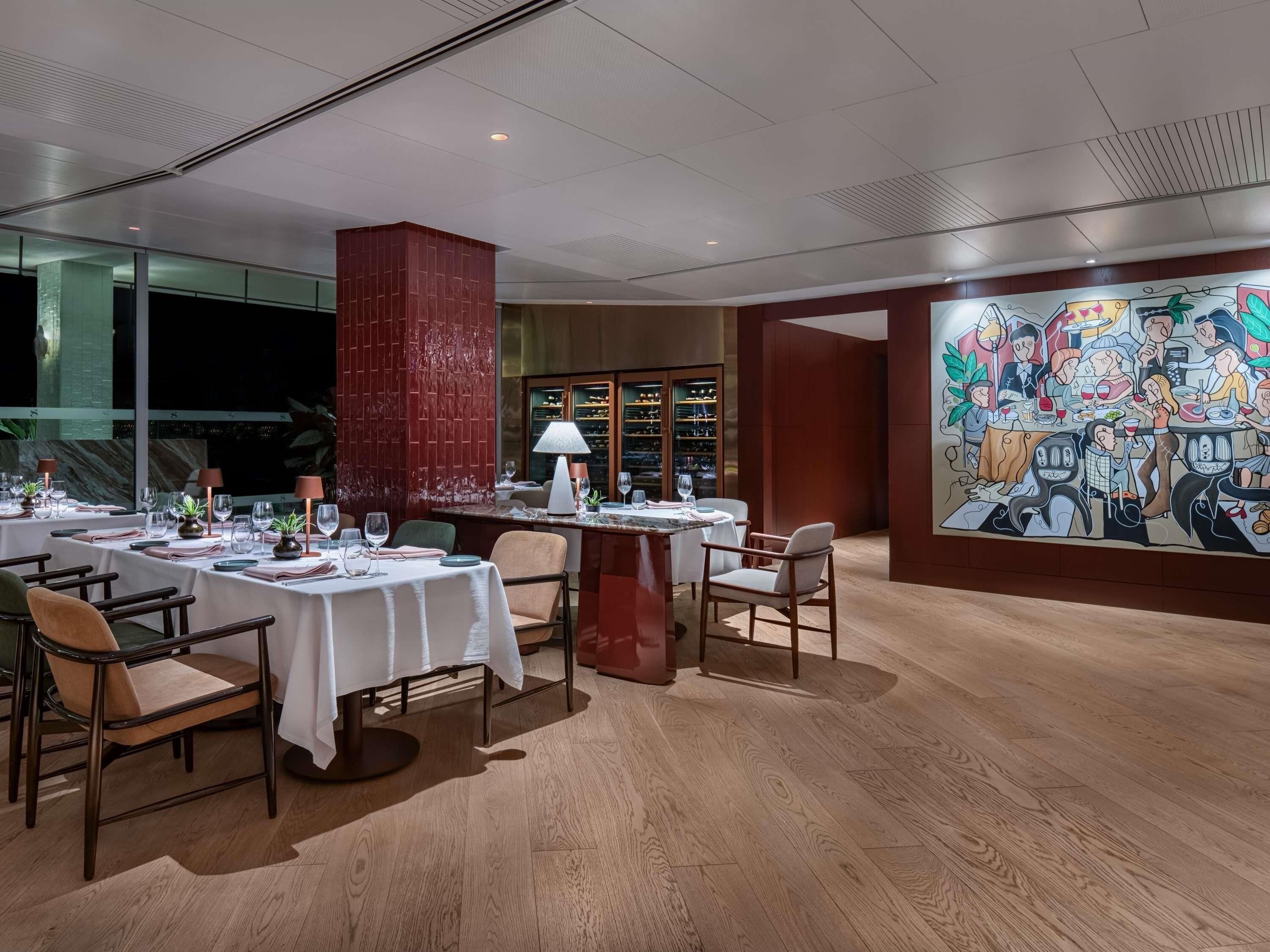 A dining area with tables set for multiple guests, a large mural on the wall, and a wine rack in the background.