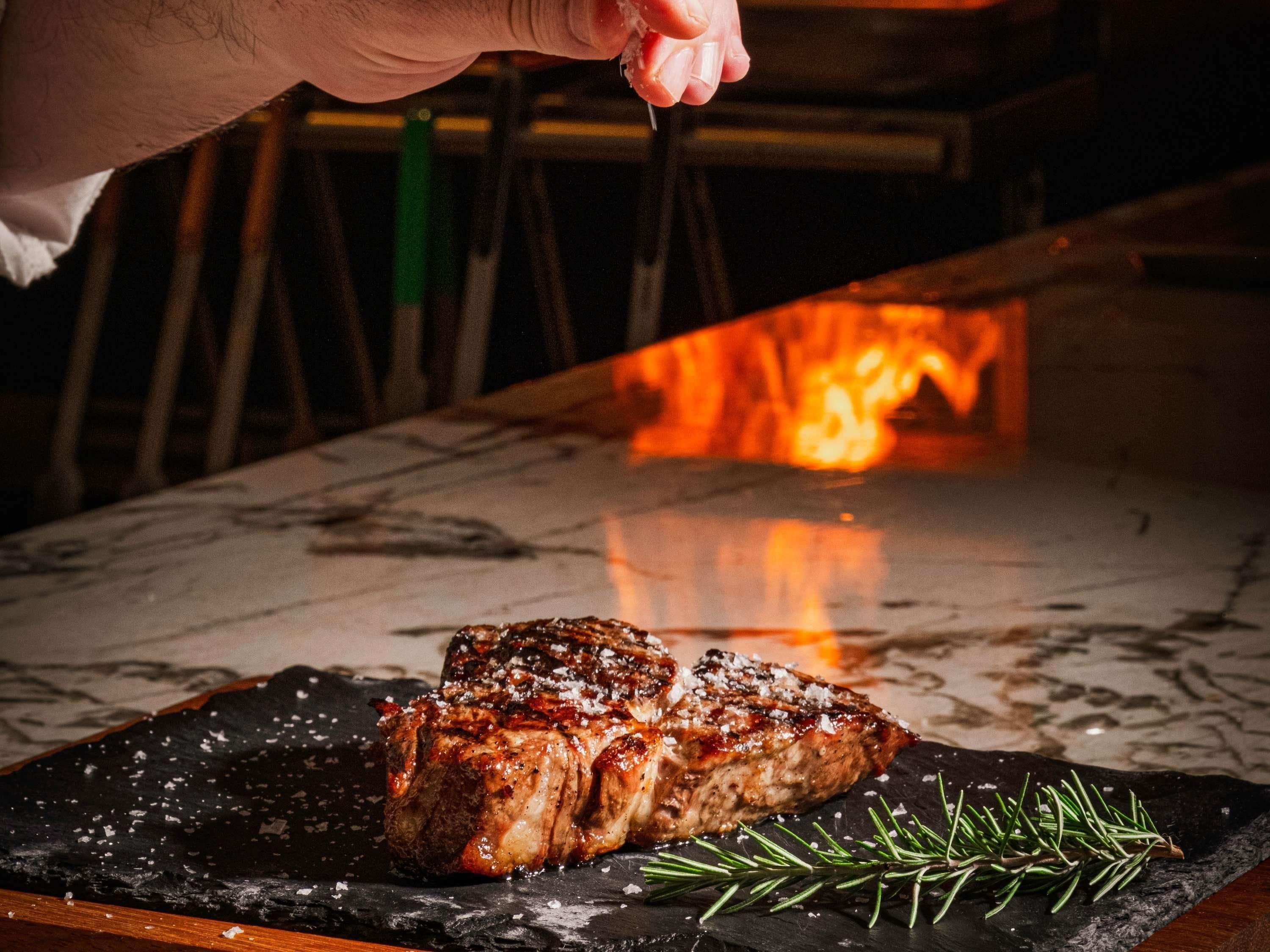 A steak being grilled over an open flame, with a sprig of rosemary on the plate.