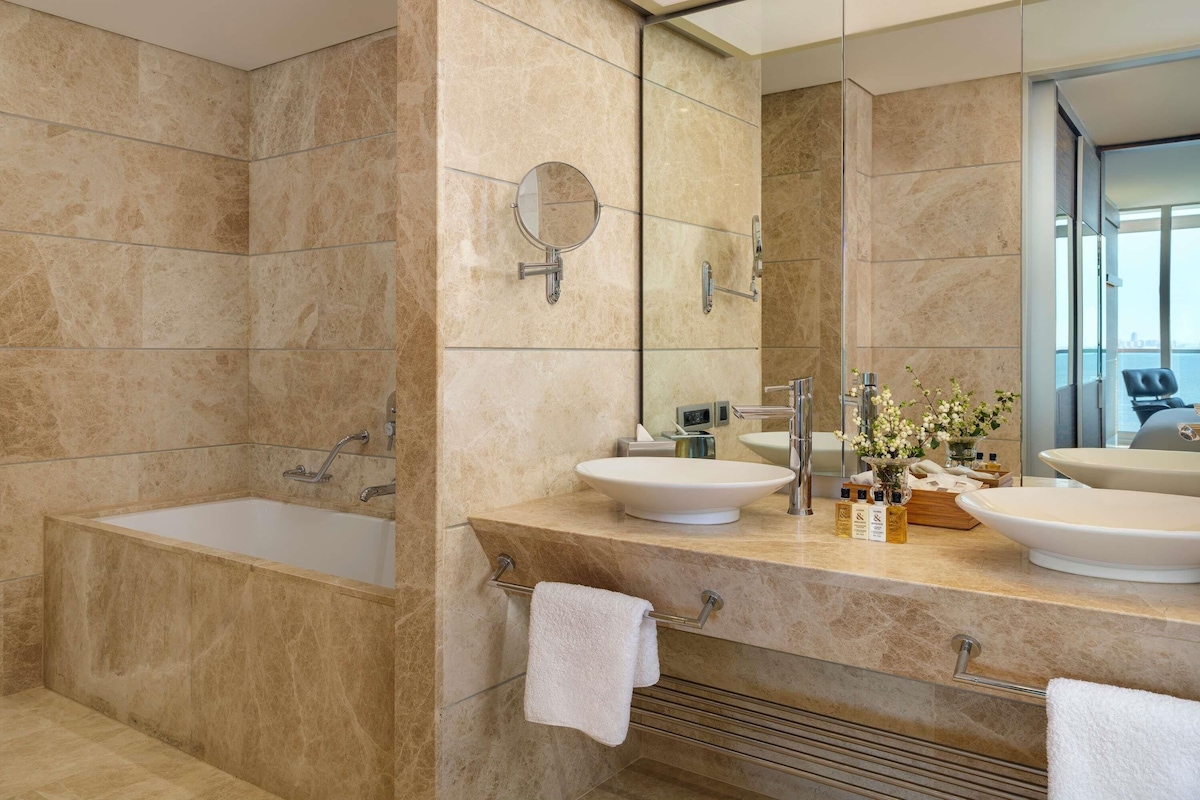 Een moderne badkamer met een badkuip, twee wastafels, een grote spiegel en uitzicht op de stad.