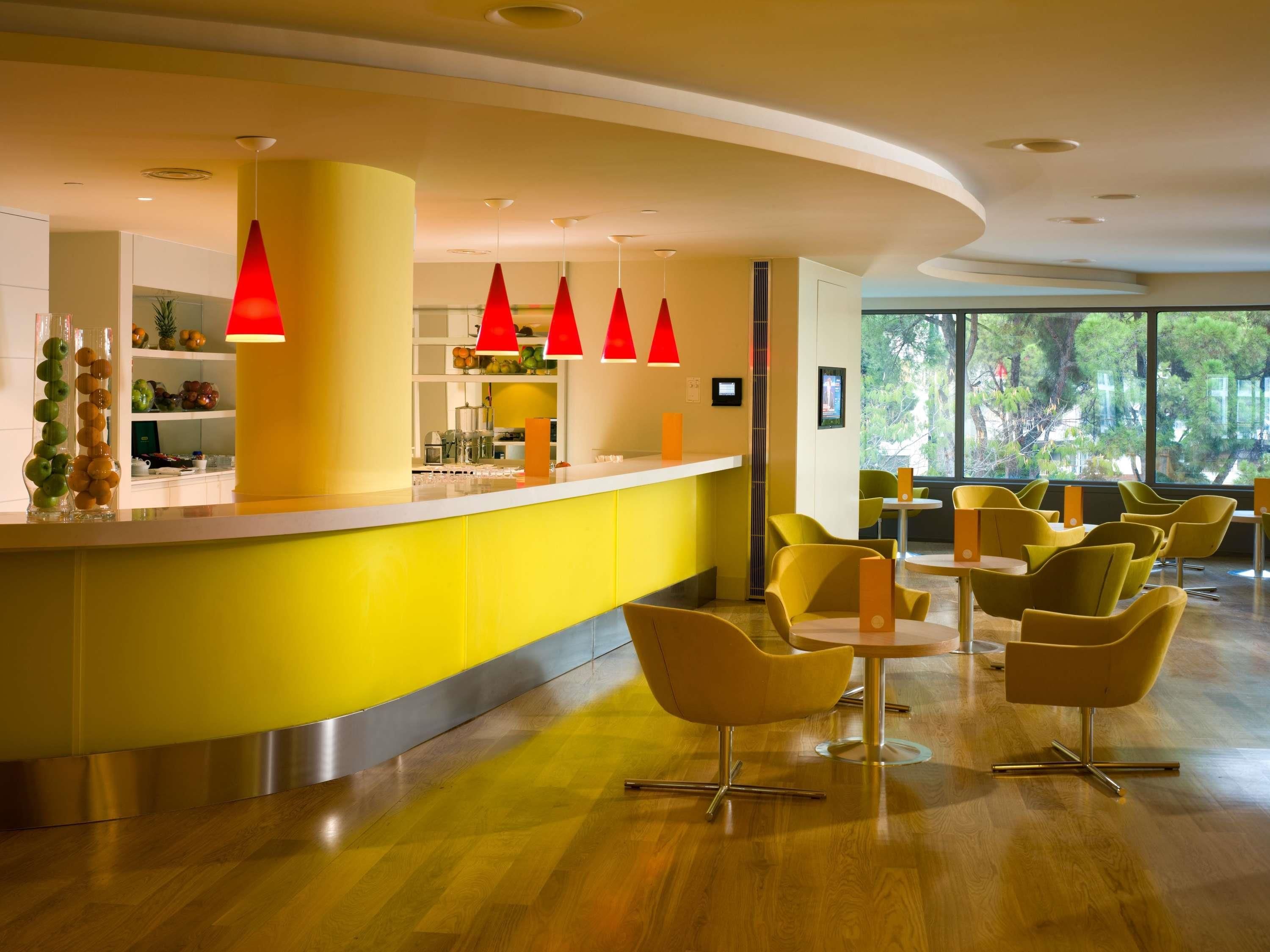 Un bar moderno con detalles amarillos, luces colgantes rojas y vistas al exterior a través de amplios ventanales.