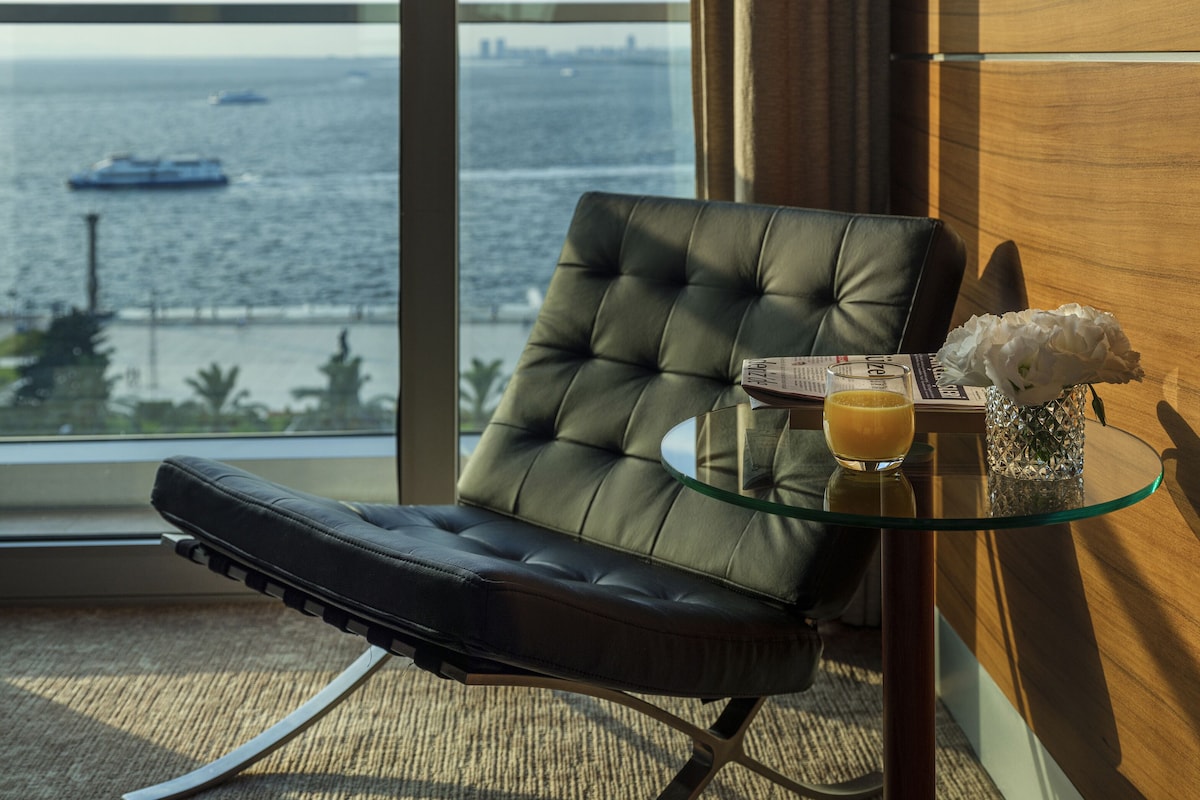 A modern room with a view of the sea, a leather chair, a glass table with a drink and flowers, and a city skyline in the background.