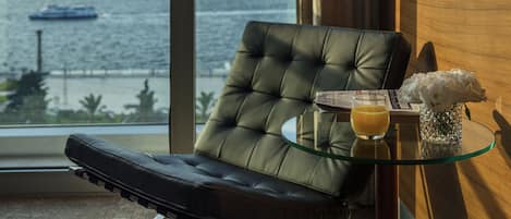 A modern room with a view of the sea, a leather chair, a glass table with a drink and flowers, and a city skyline in the background.