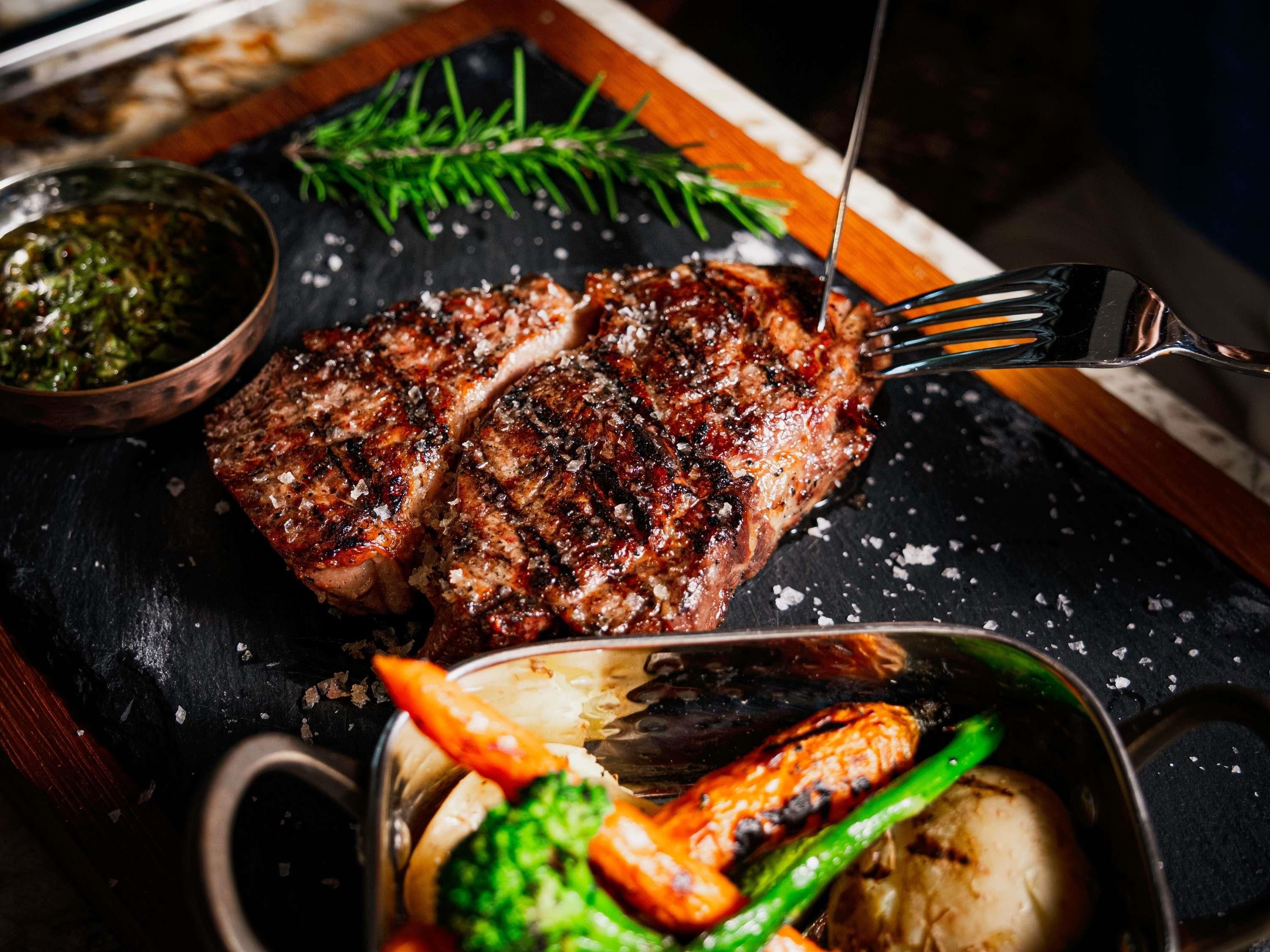 Un bistec con un tenedor, acompañado de verduras y un bol de salsa sobre una superficie de pizarra oscura.