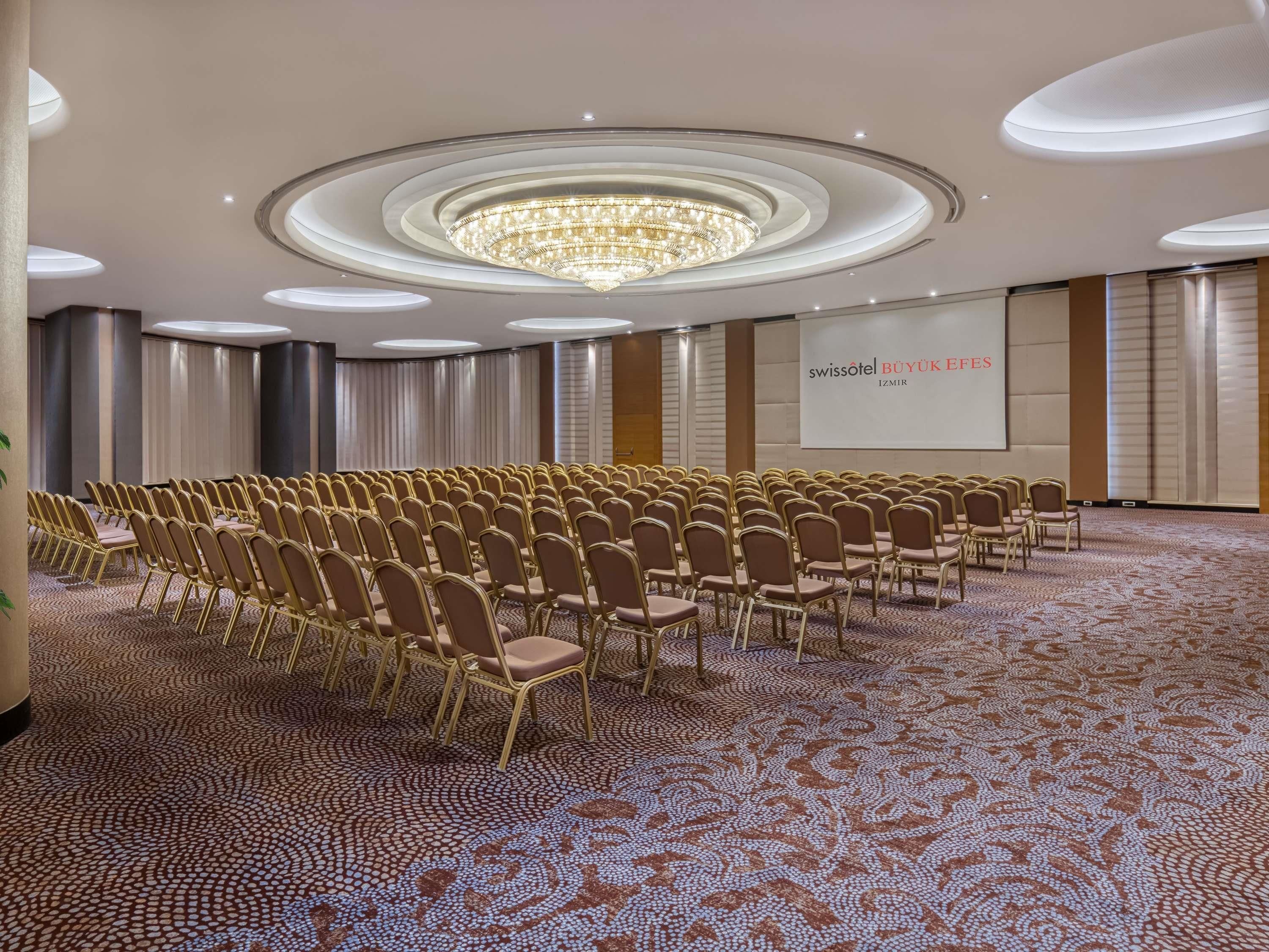 A conference room with rows of chairs, a large chandelier, and a screen displaying 'swissotel BUYUKIFESES IZMIR'.