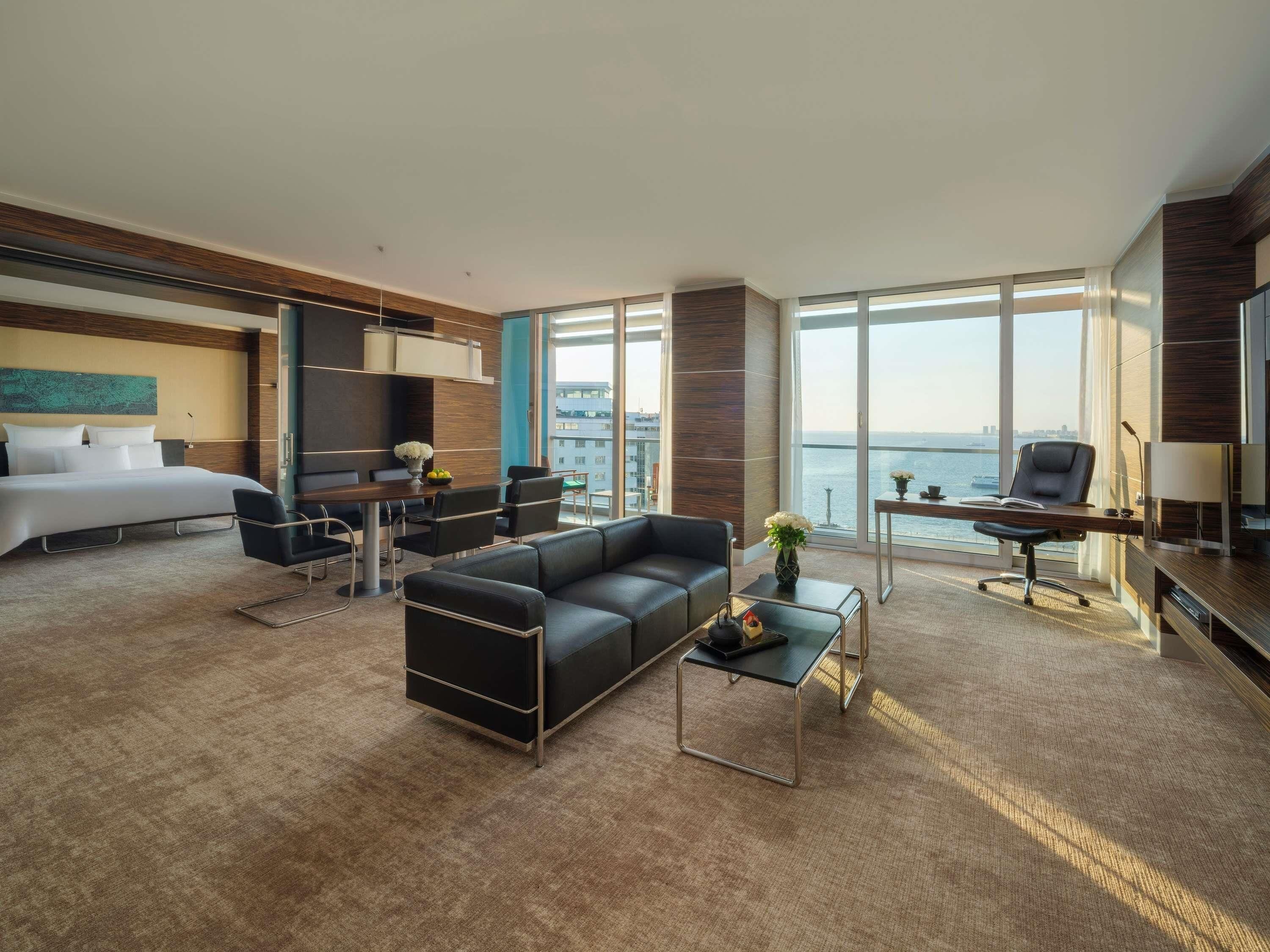 A modern hotel room with a bed, a sofa, a desk, and a view of the sea.