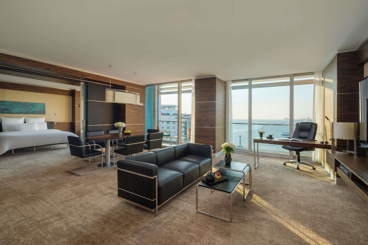 A modern hotel room with a bed, a sofa, a desk, and a view of the sea.