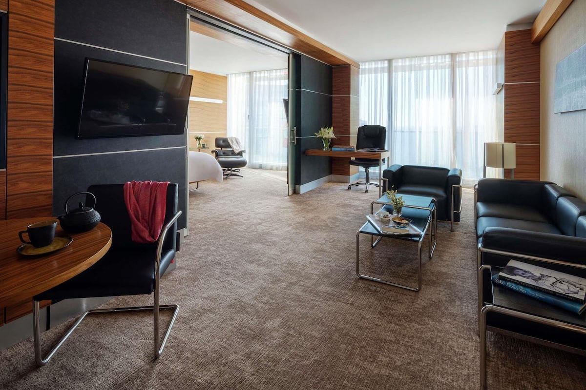 A modern hotel room with a flat-screen TV, leather seating, a coffee table, and a desk with a chair.