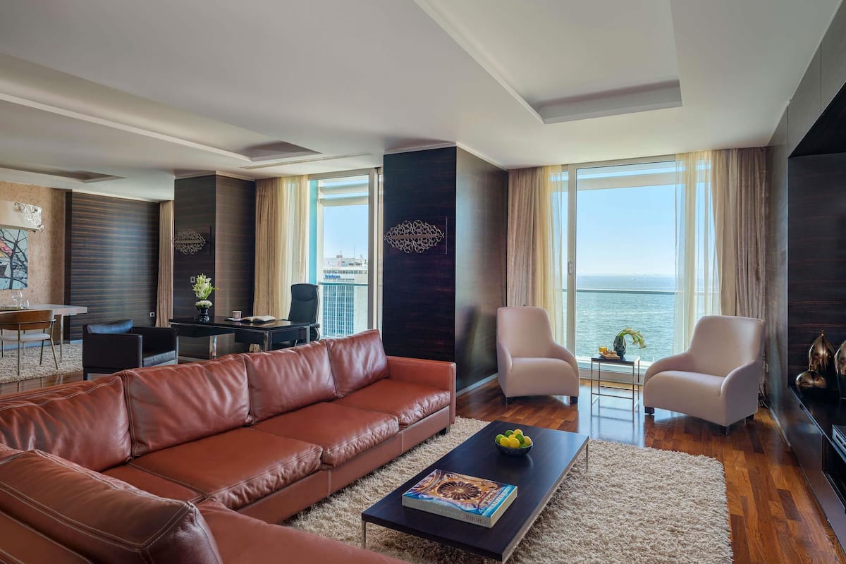 Una sala de estar moderna con un sofá de cuero, una mesa de centro y dos sillones. Se observa un comedor con una mesa y sillas. Grandes ventanales ofrecen vista al mar.
