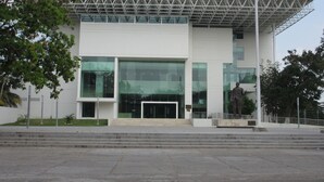 Un edificio moderno con grandes ventanales, estructura de techo metálica y una estatua en primer plano.