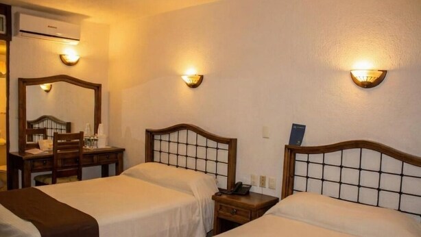 Habitación de hotel con dos camas, un escritorio con silla y un espejo. Hay luces empotradas en la pared y un equipo de aire acondicionado.