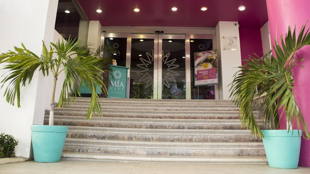 A hotel entrance with a staircase leading up to a glass door, potted plants on either side, and a pink wall to the right. - Mia City Villahermosa (Villahermosa)