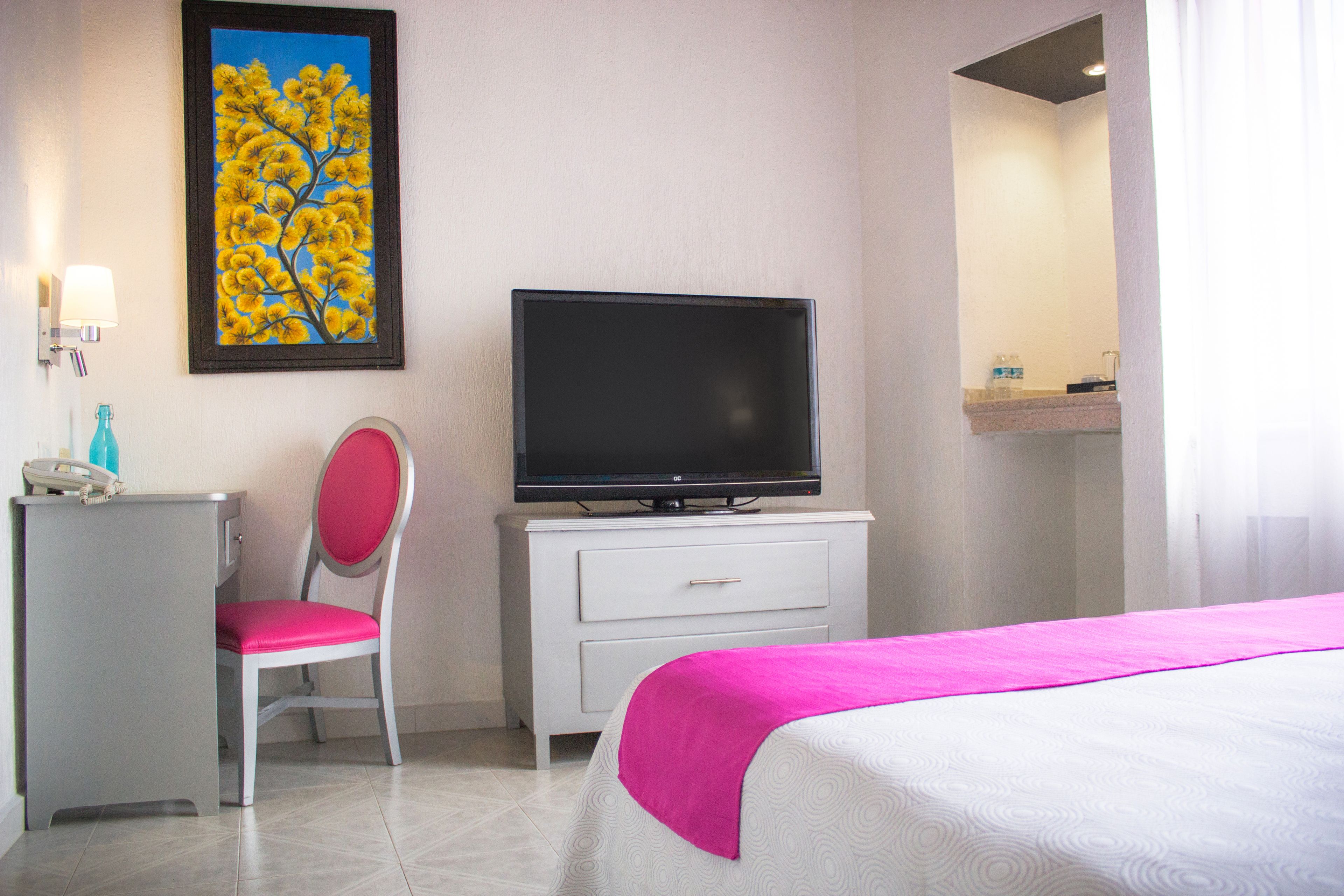 Habitación de hotel con cama, televisión sobre un tocador blanco, una silla, escritorio y un cuadro en la pared.