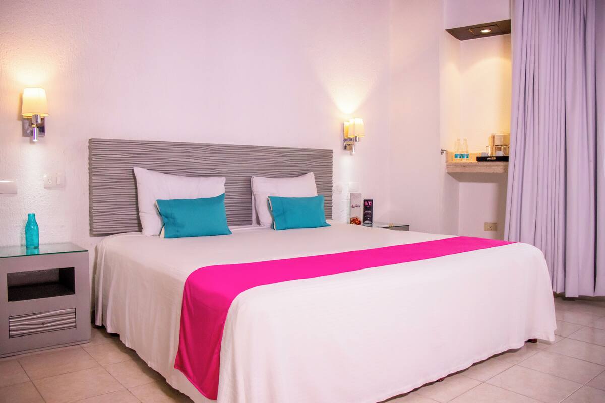 A hotel room with a large bed, two bedside tables, a nightstand, a lamp, and a shelf with toiletries.
