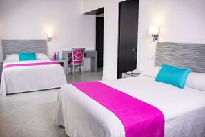 A hotel room with two beds, each with a white bedspread and a bright pink accent. There is a desk with a chair, a nightstand with a lamp, and a door in the background.