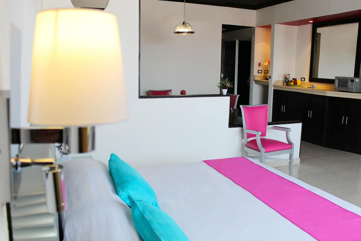 A hotel room with a bed, a chair, a desk, a lamp, and a kitchen area.