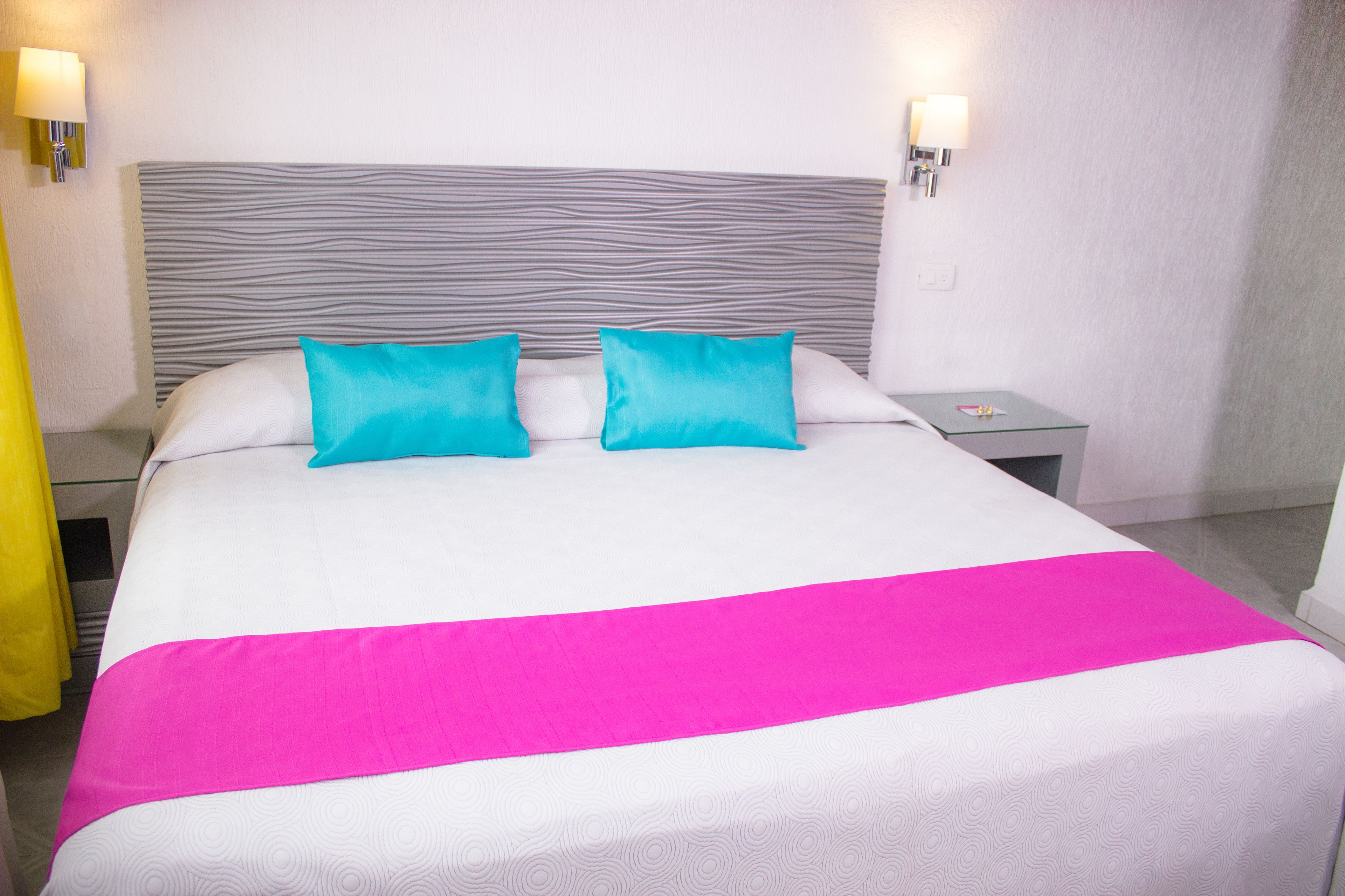 Una habitación de hotel con una cama, dos almohadas, una mesita de noche y dos lámparas fijadas a la pared.