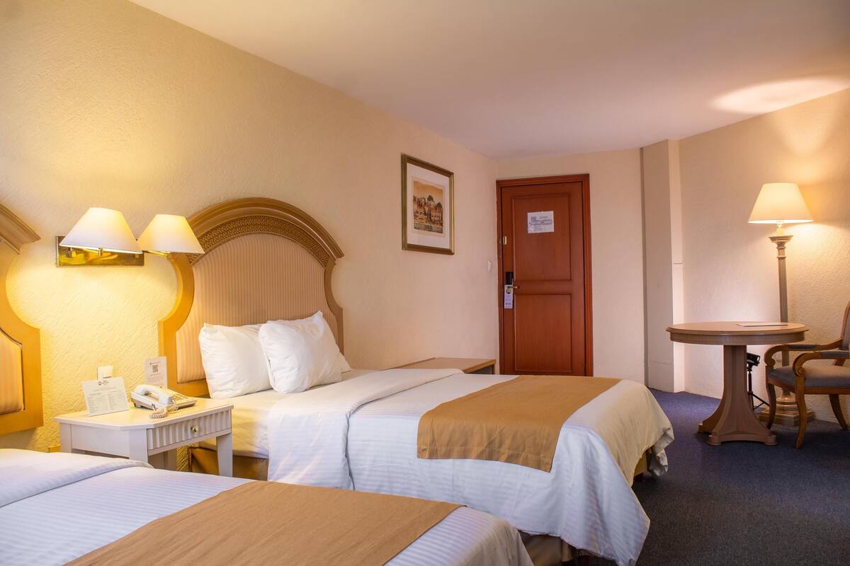 Une chambre d’hôtel avec deux lits, une tête de lit en bois, une petite table avec une chaise et une lampe.