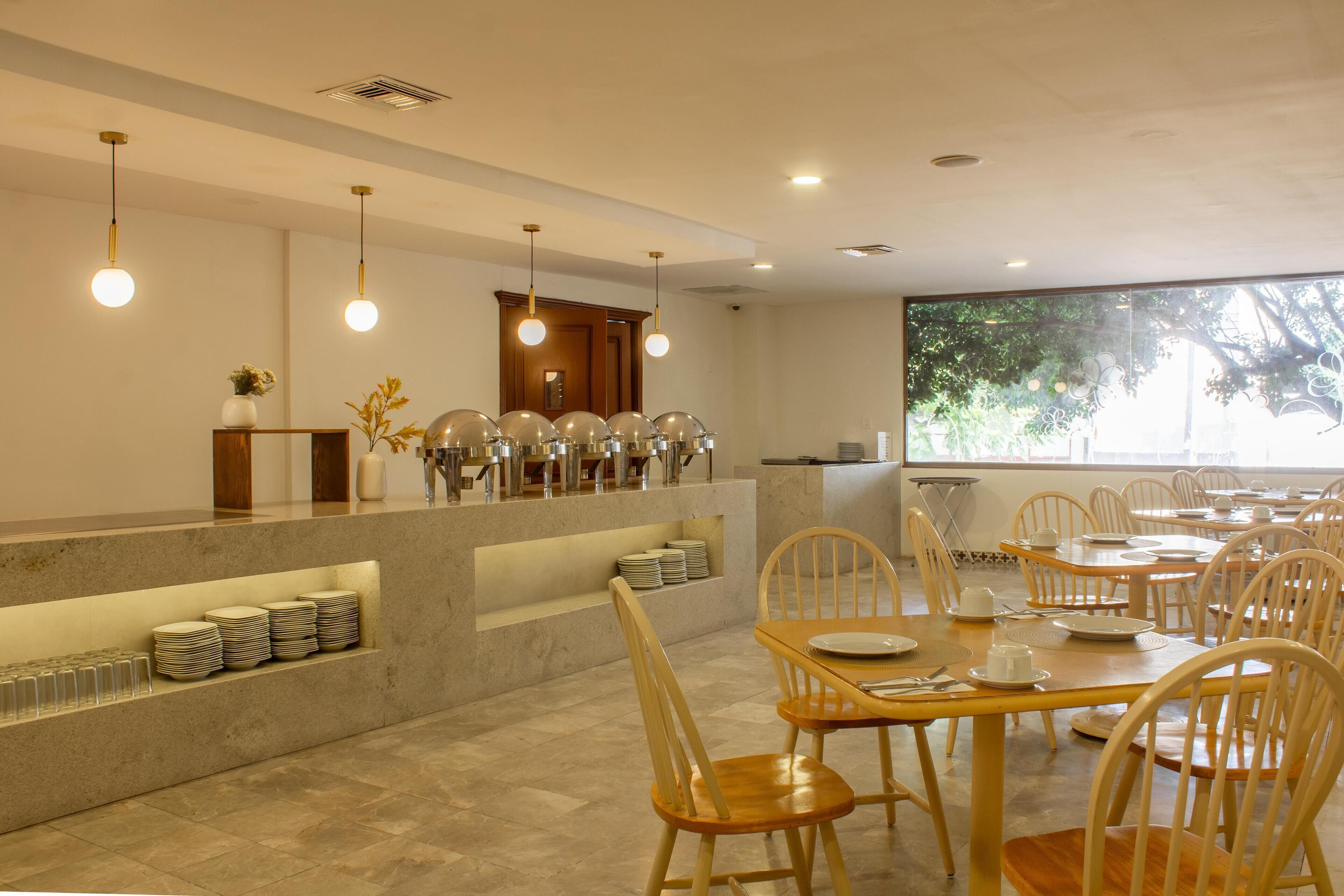 A dining area with multiple tables and chairs, a buffet counter with stacked plates and glasses, and pendant lighting.