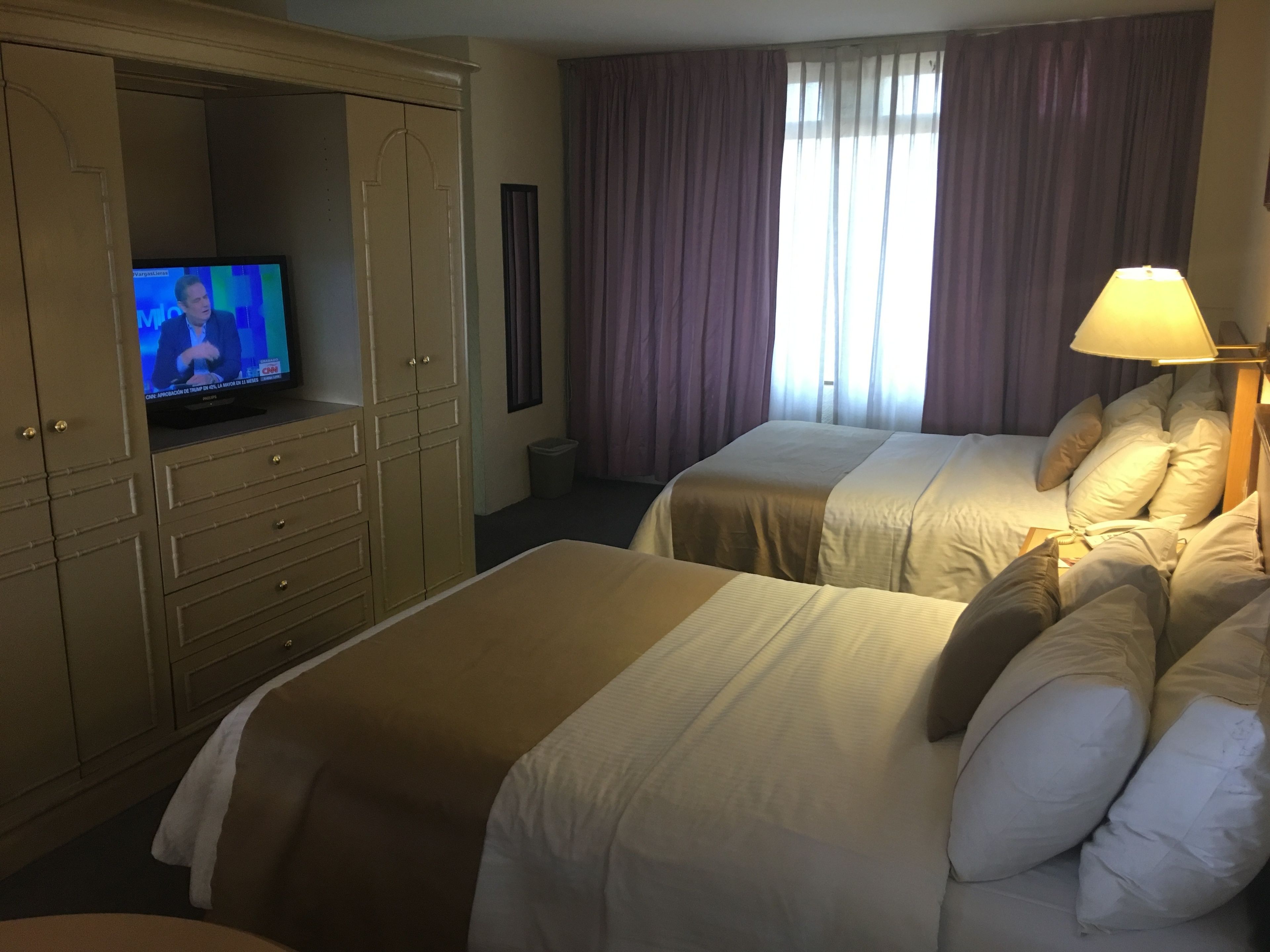Quarto de hotel com duas camas, televisão em um móvel, uma luminária e uma janela com cortinas.