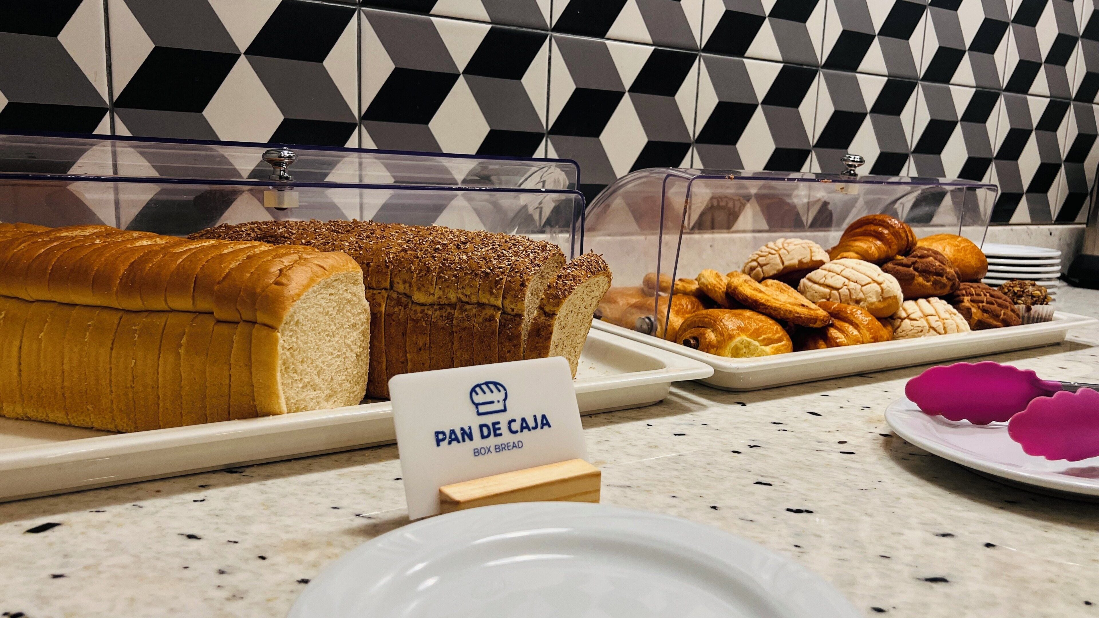 A bakery display with various breads and pastries, a sign indicating 'PAN DE CAJA', and a tiled backsplash.