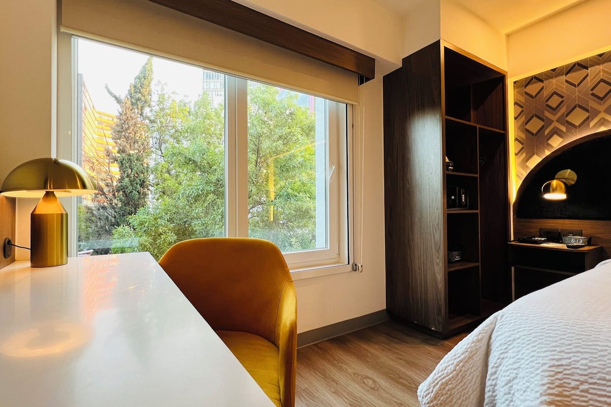 Habitación de hotel con cama, escritorio con lámpara, silla, armario y ventana con vistas a árboles.