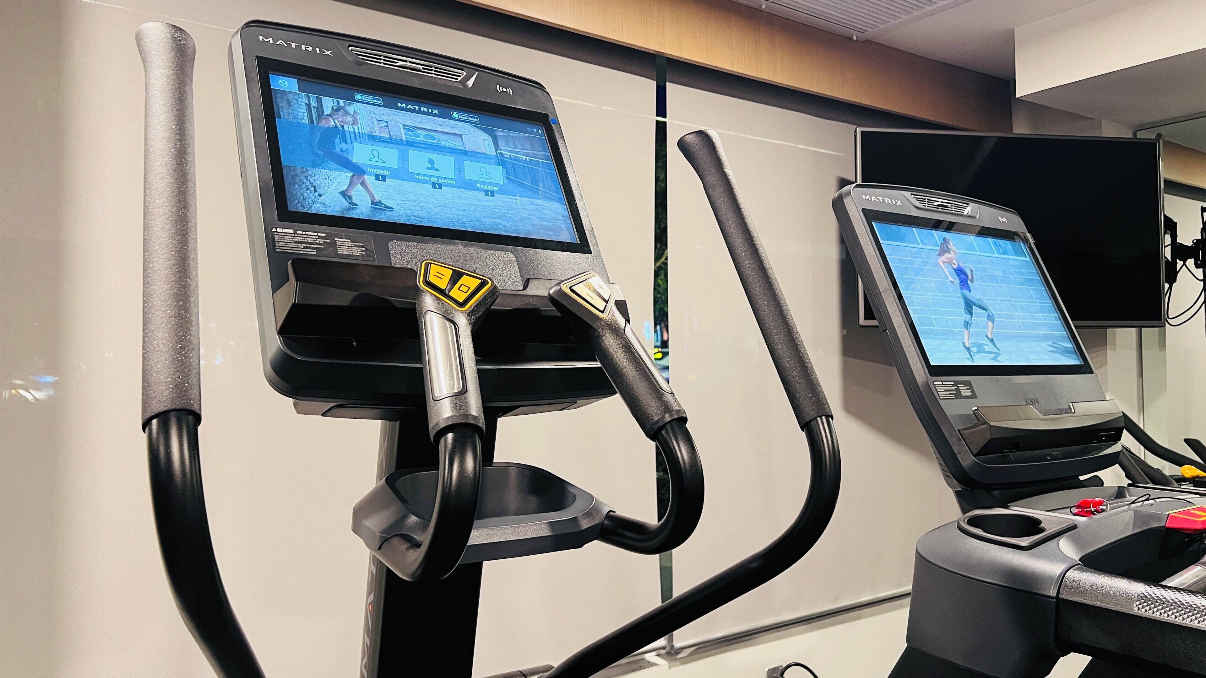 Two stationary exercise bikes with digital screens displaying workout programs, connected to a wall-mounted control panel.