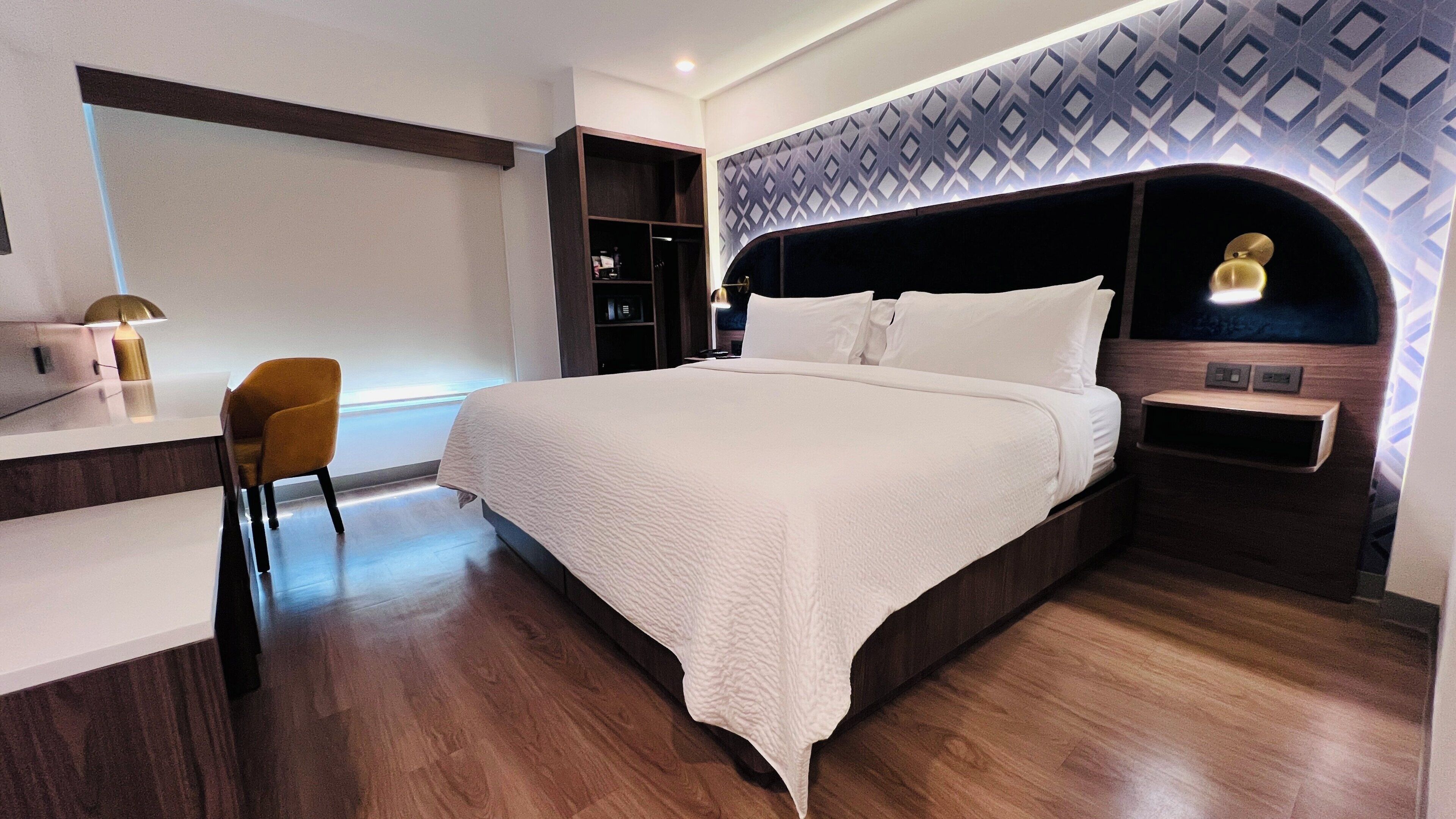 Una habitación de hotel moderna con una cama grande, un escritorio con lámpara, una silla y un papel tapiz estampado.