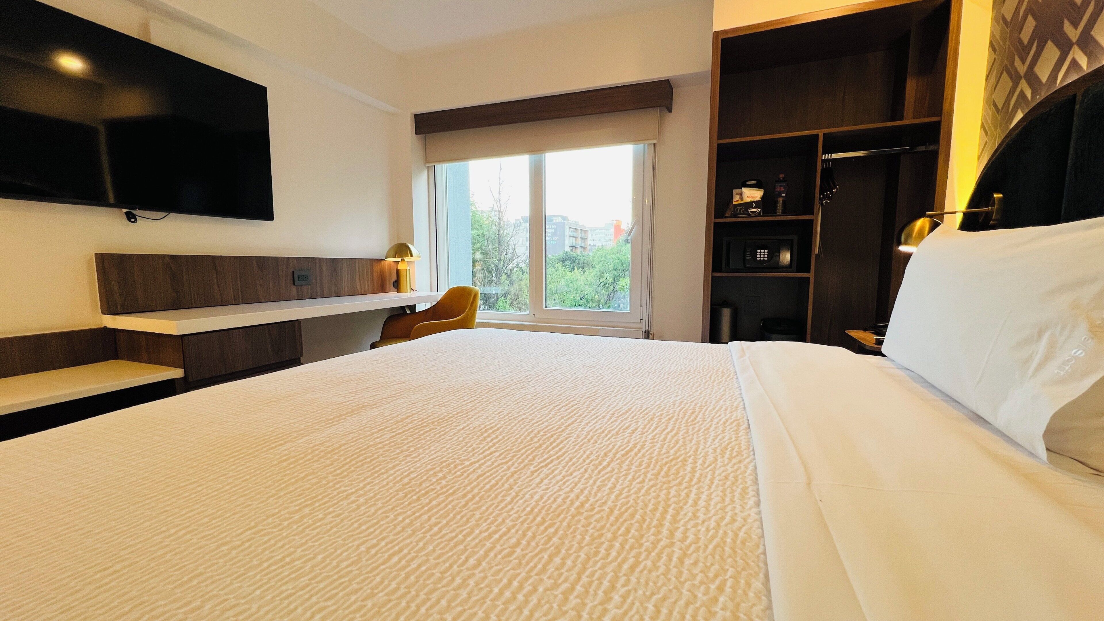 Une chambre d  ’hôtel avec un grand lit, un téléviseur fixé au mur, une fenêtre donnant sur des immeubles, une armoire et une petite table avec une lampe.