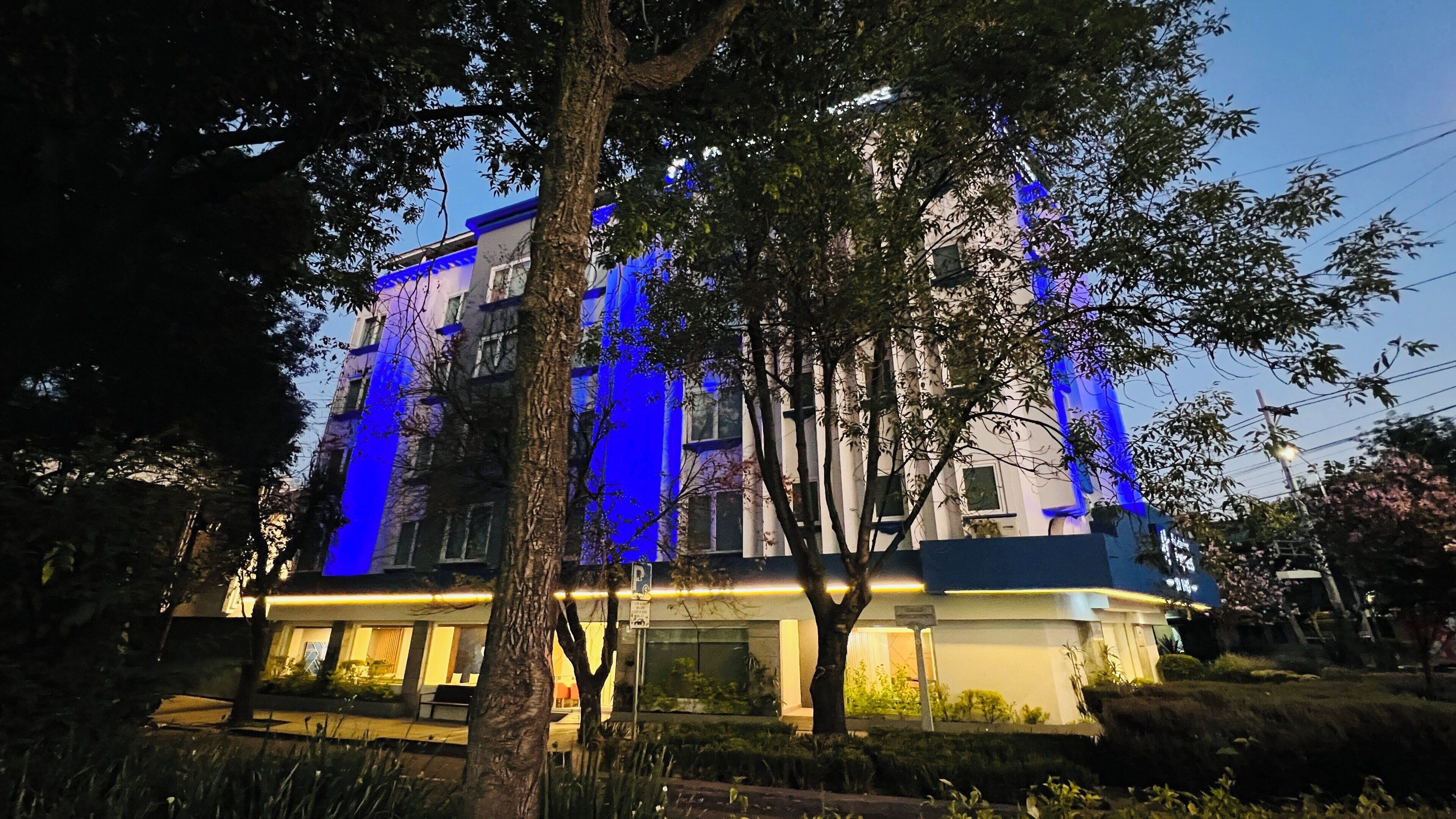 Un immeuble à plusieurs étages dont la façade est illuminée d’une lumière bleue, entouré d’arbres et d’une pelouse bien entretenue.
