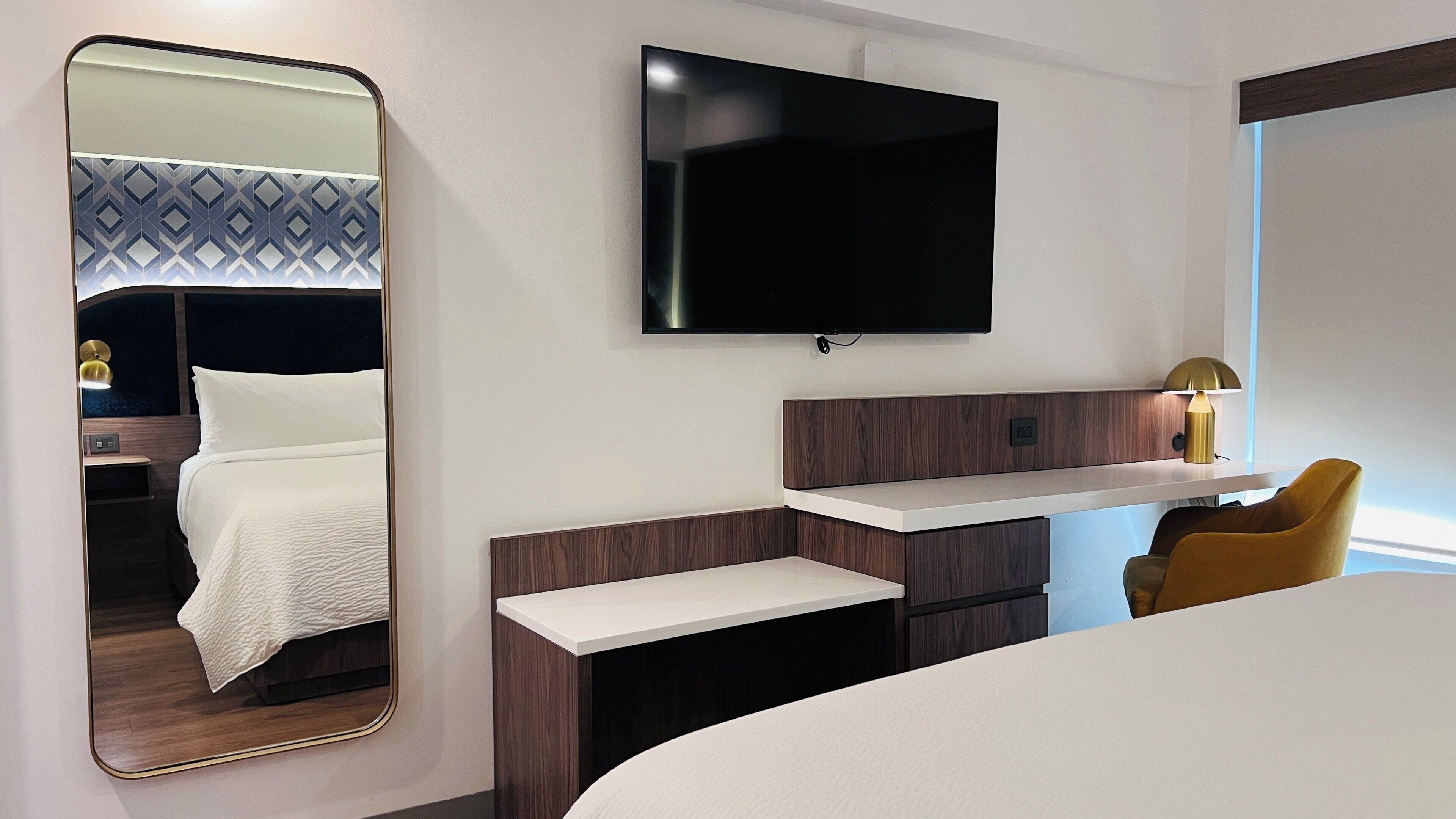 Een moderne hotelkamer met een groot bed, nachtkastje, een aan de muur bevestigde televisie en een spiegel die de kamer weerkaatst.