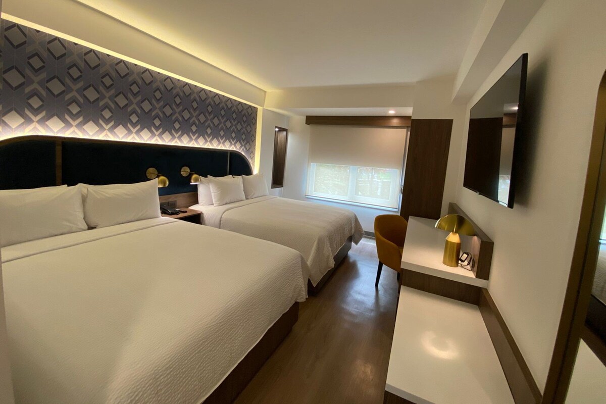 Habitación de hotel con dos camas, televisor, escritorio con lámpara y pared con diseño.