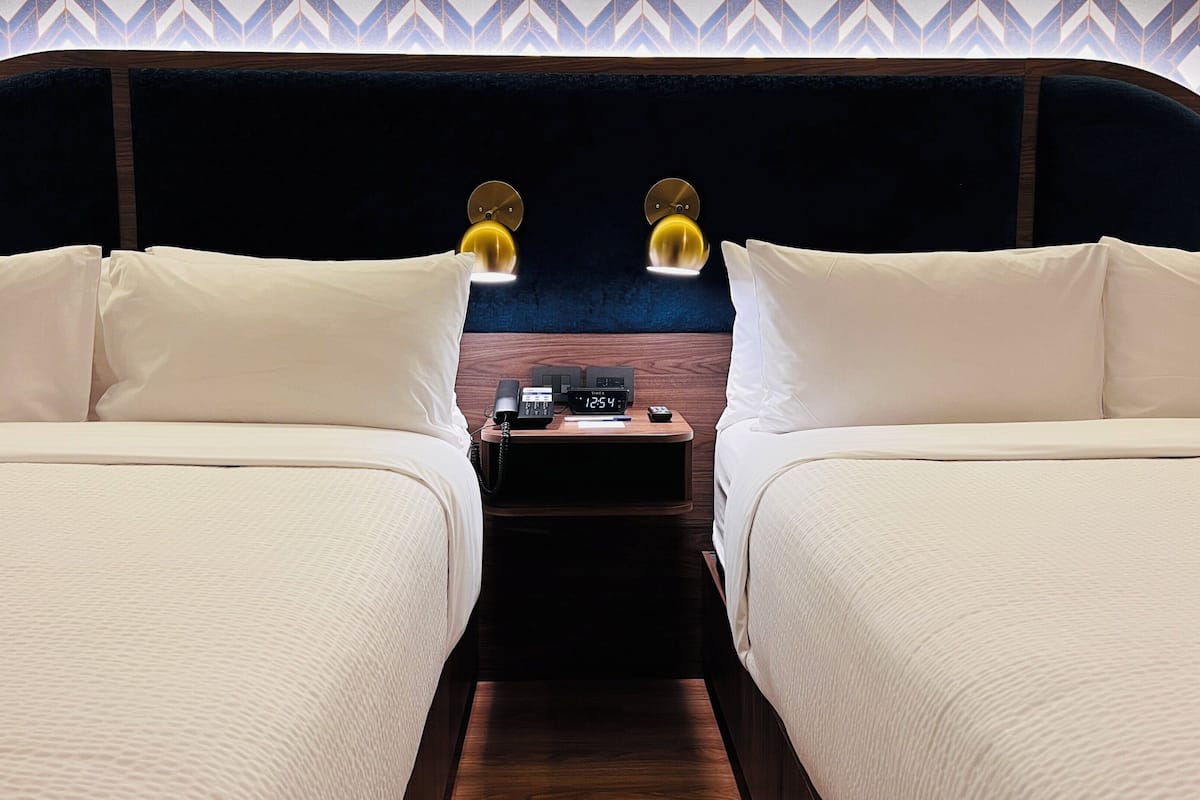 Habitación de hotel con dos camas, cabecera de madera, mesita de noche con un reloj y un papel tapiz estampado.